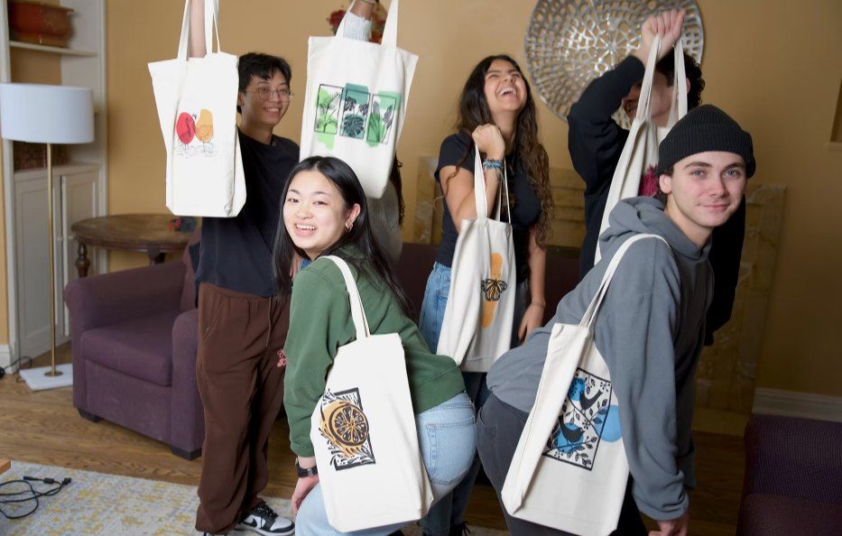 Six friends laughing and holding up Angaza Taa totes in a living room