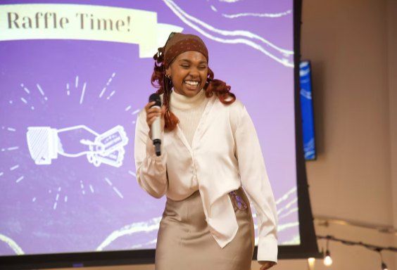 Ally Karabu smiling and speaking into a microphone in front of a Raffle Time slide