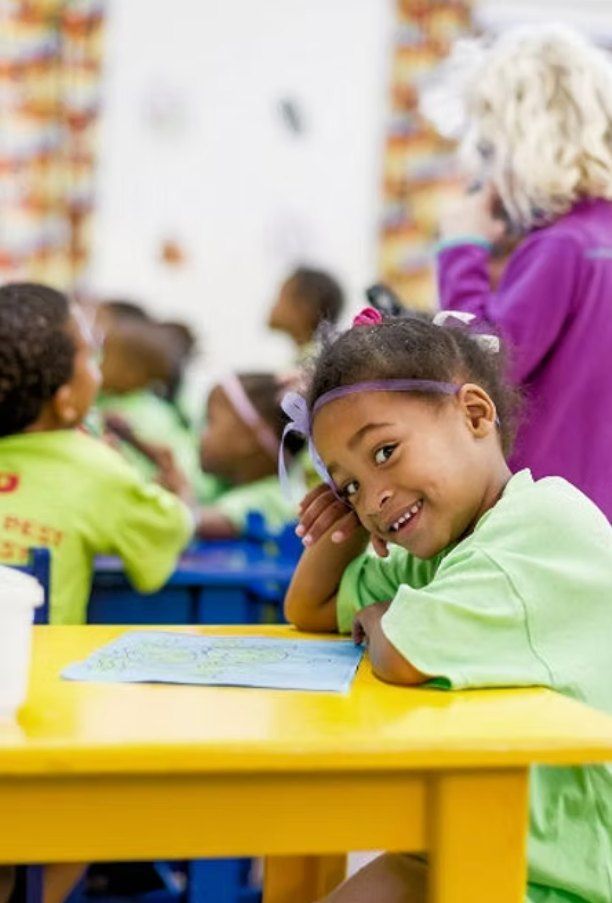 A smiling young girl in a green shirt at a yellow classroom table