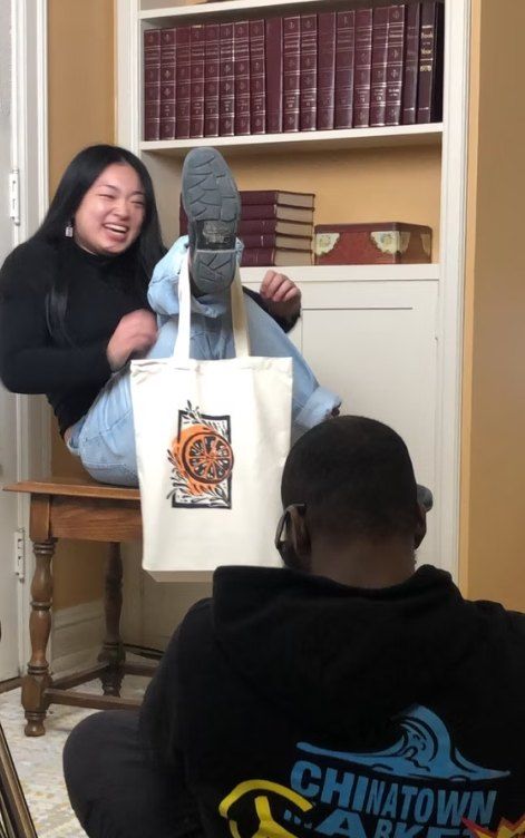 A laughing woman holds up an Angaza Taa tote with an orange citrus print while a friend photographs her